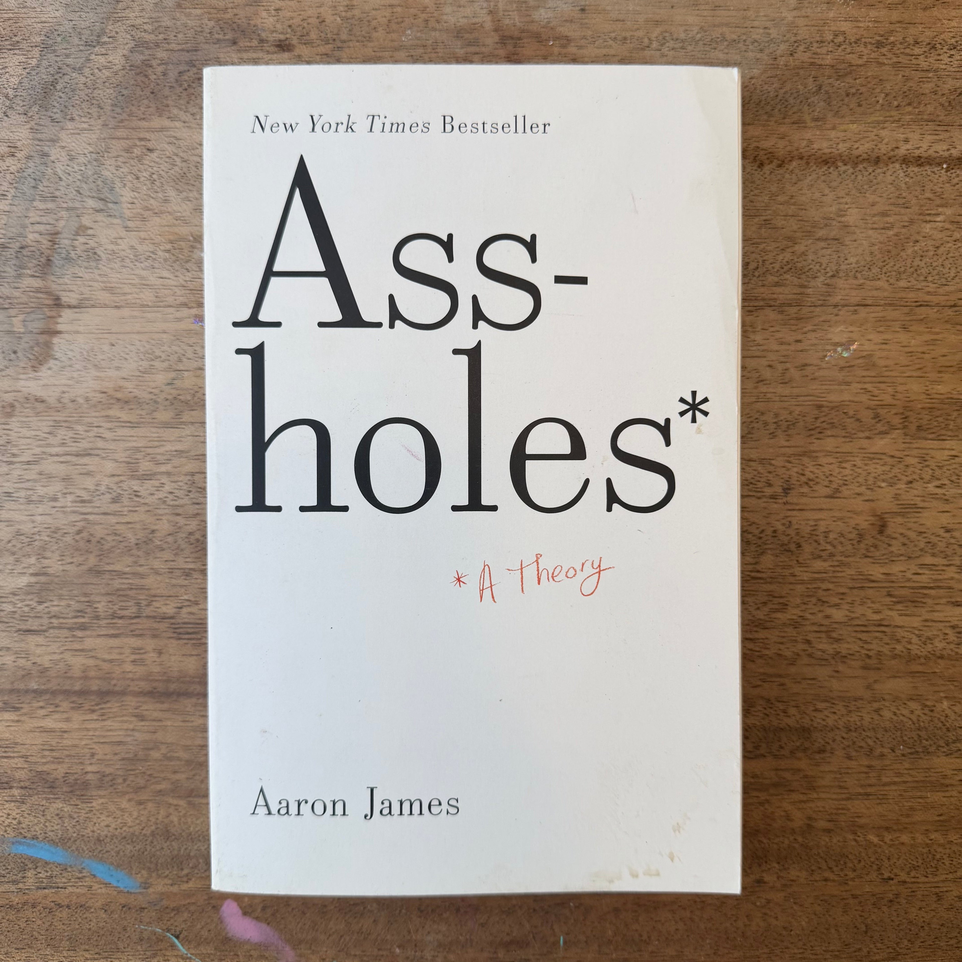 The book cover of 'Ass. Holes' by Aaron James, featuring a cream-colored background with bold black typography and a small red handwritten star annotation, photographed flat on a wooden surface.