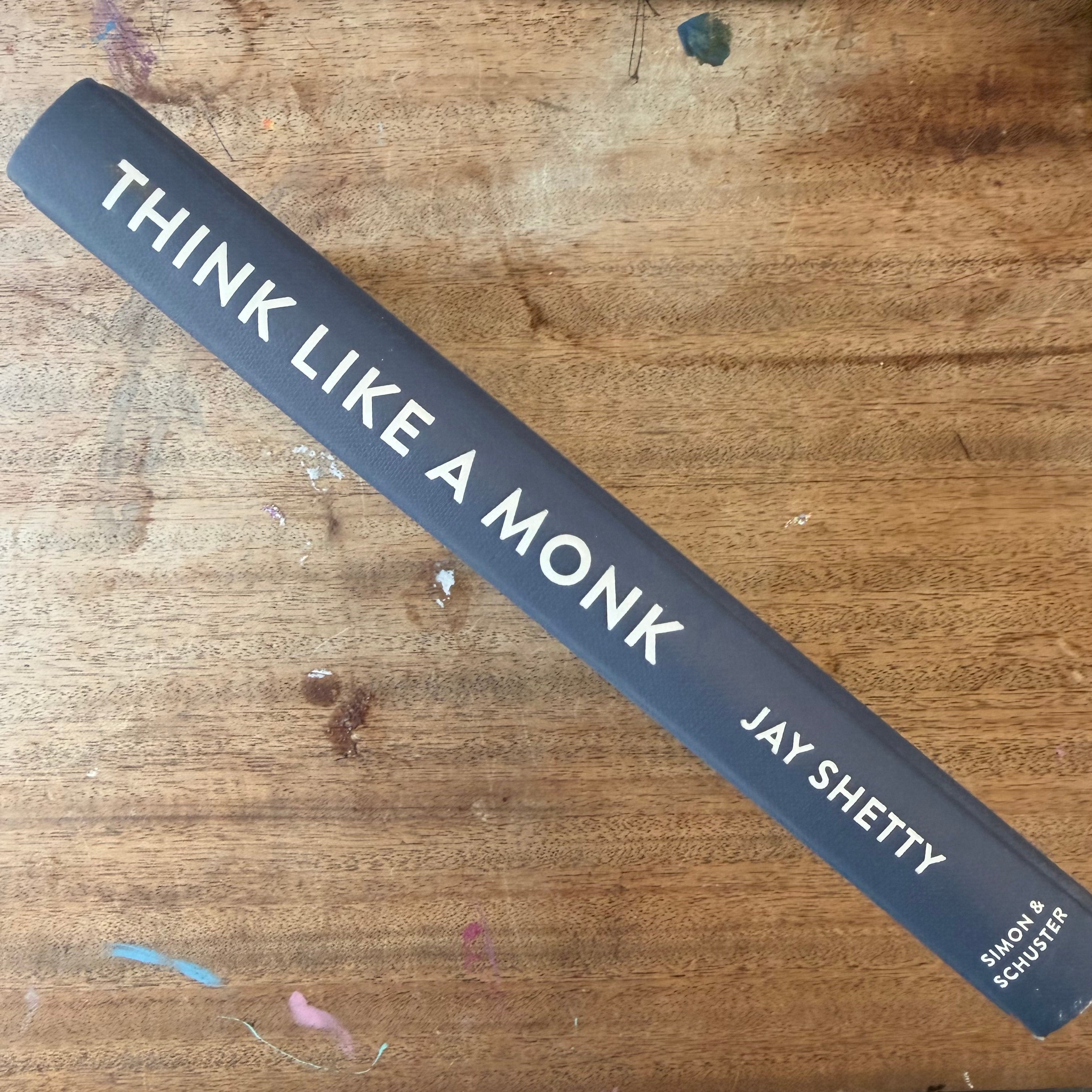 The spine of the book 'Think Like a Monk' by Jay Shetty, published by Simon & Schuster, displayed diagonally on a rustic wooden table.
