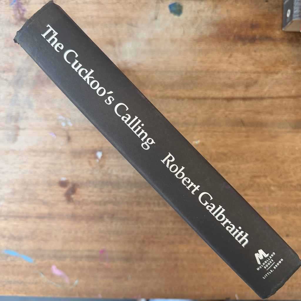The spine of a hardcover book titled 'The Cuckoo's Calling' by Robert Galbraith, featuring a dark brown cover with white text, placed diagonally on a wooden surface.