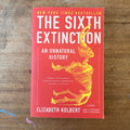 The book 'The Sixth Extinction' by Elizabeth Kolbert, featuring a bright red cover with a yellow dinosaur skeleton illustration and bold white and yellow text, placed on a wooden surface.