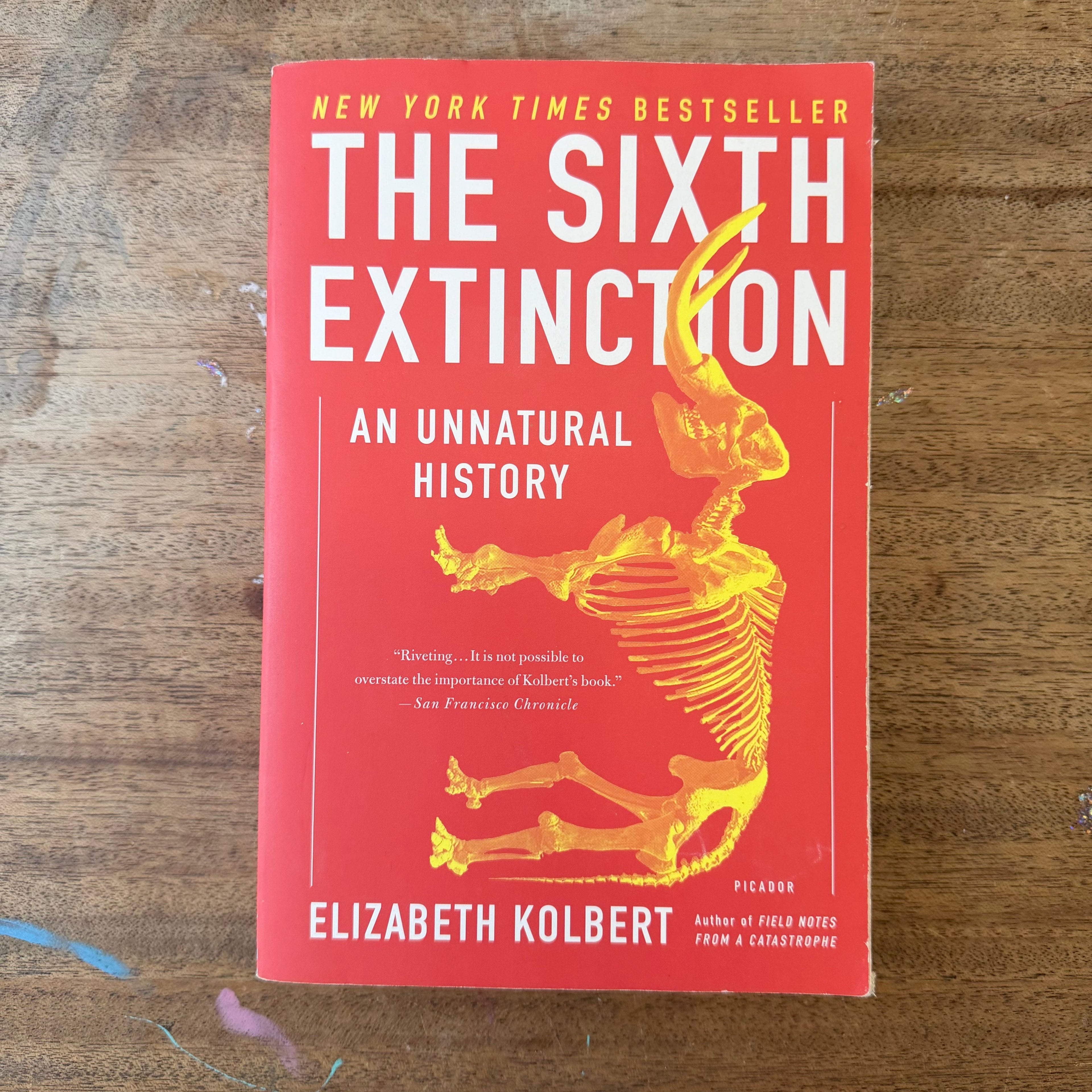 The book 'The Sixth Extinction' by Elizabeth Kolbert, featuring a bright red cover with a yellow dinosaur skeleton illustration and bold white and yellow text, placed on a wooden surface.