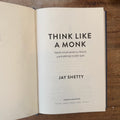 An open book titled 'Think Like a Monk' by Jay Shetty, showing the title page on a wooden surface.
